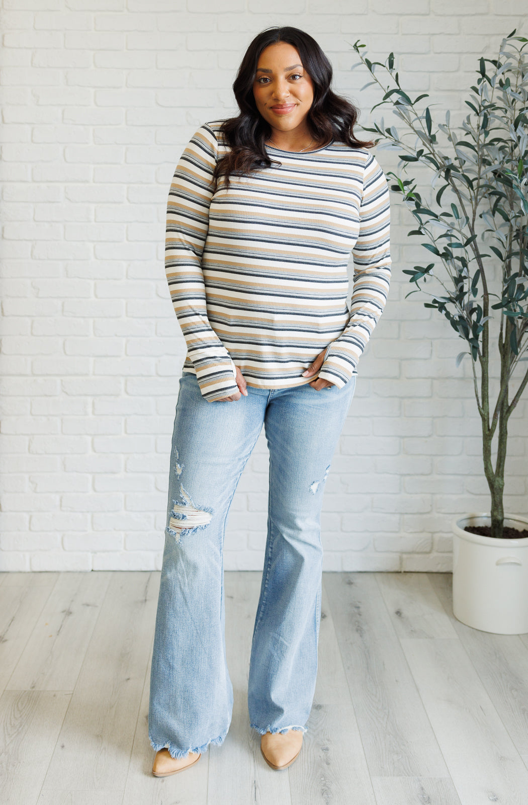 Woman wearing a striped sweater and jeans standing in a room with a white brick wall and plant.