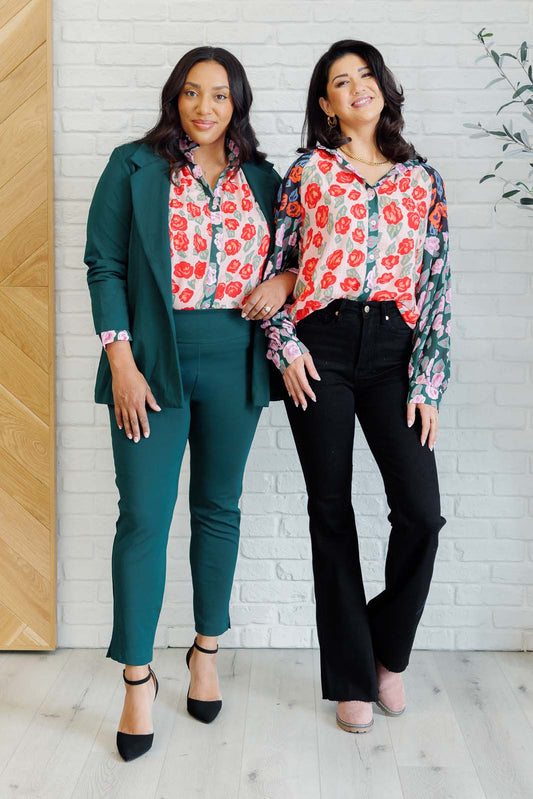 Two women wearing floral tops and pants standing against a white brick wall.
