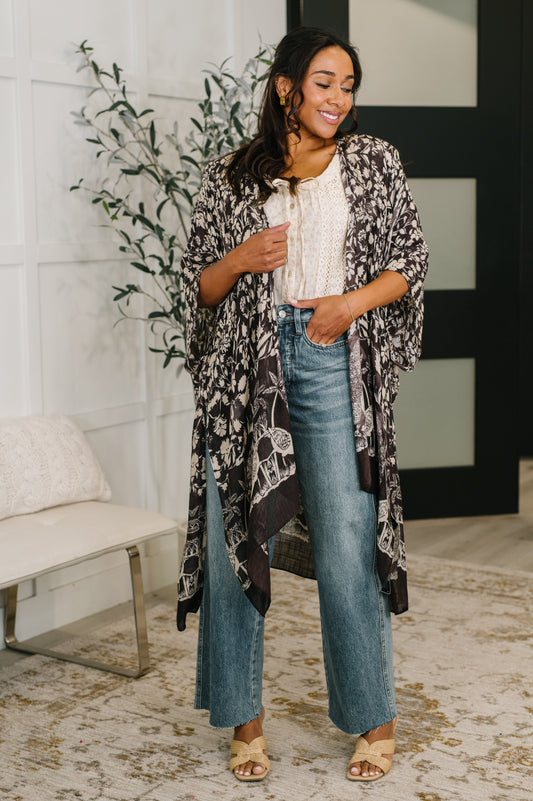 Woman wearing a patterned kimono over a white top and blue jeans in a room with a plant and bench.