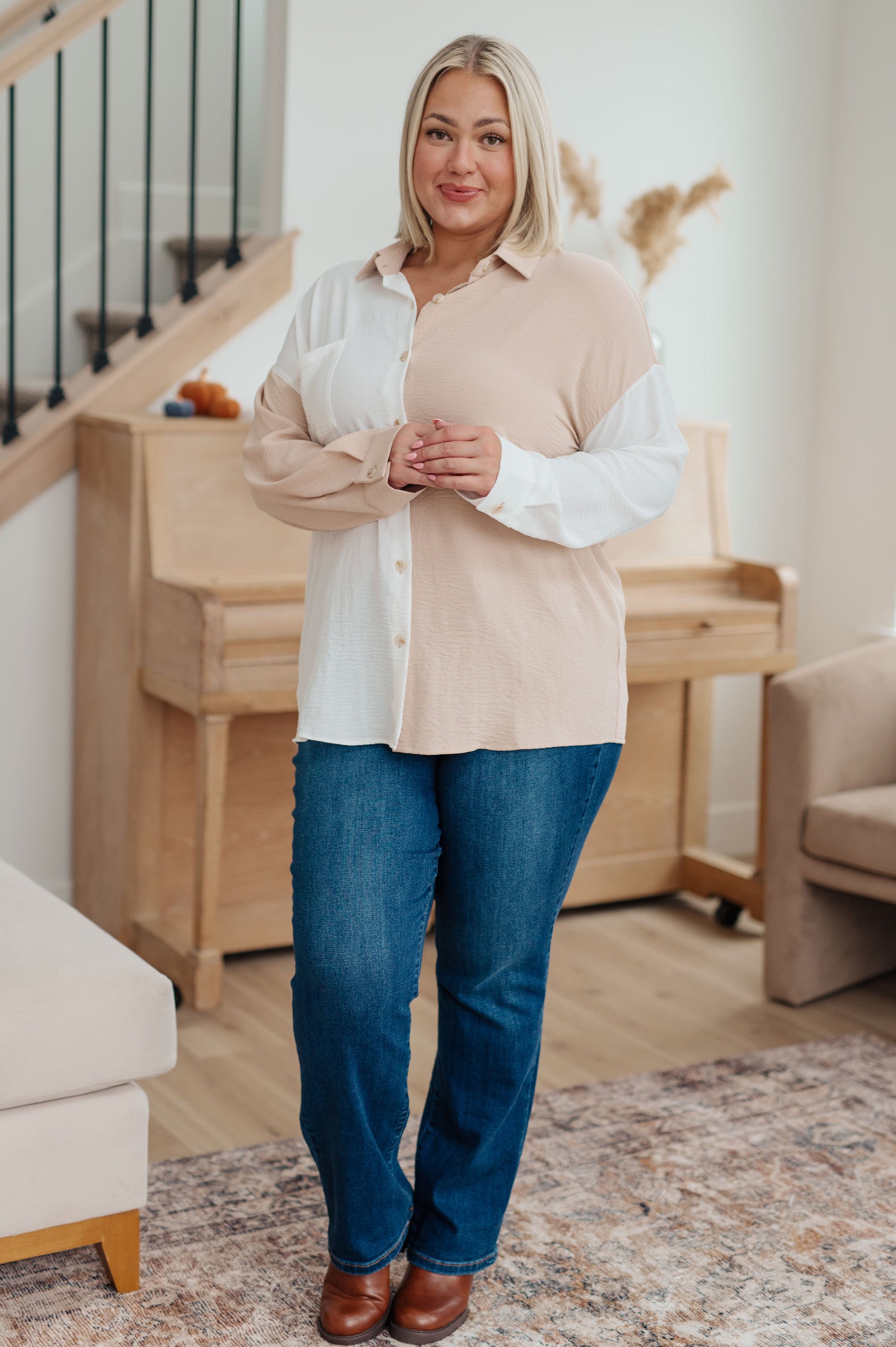 Woman standing in a living room wearing a beige shirt over a white blouse and blue jeans.