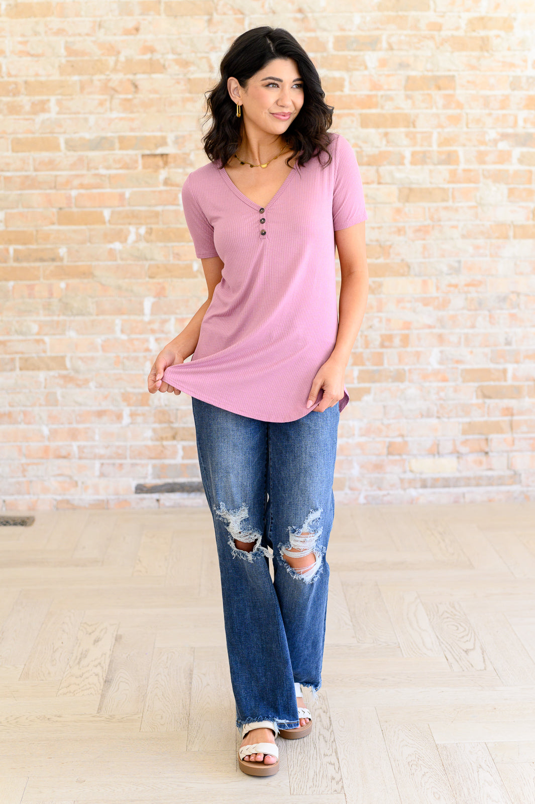 Woman wearing a pink top and blue jeans standing against a brick wall.
