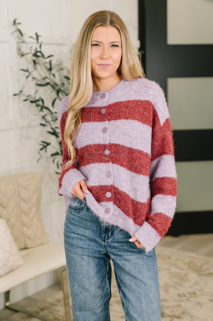 A woman wearing a fuzzy knit cardigan with horizontal stripes in dusty pink and terracotta red, paired with blue jeans, standing in a brightly lit indoor setting.