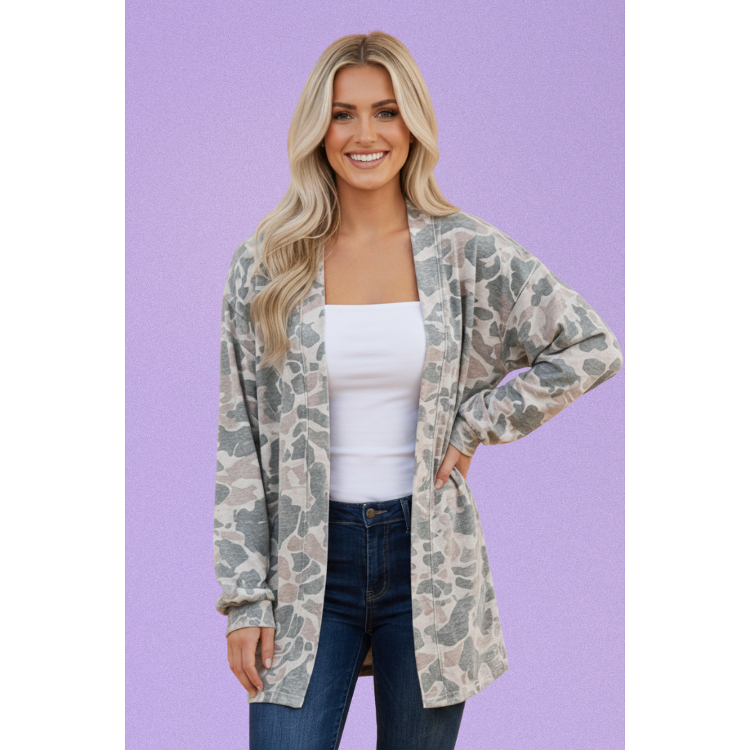 A woman modeling a lightweight open-front cardigan with a muted camouflage pattern in beige, gray, and olive green, worn over a white tank top and blue jeans against a solid lavender background.