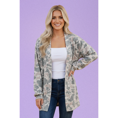 A woman modeling a lightweight open-front cardigan with a muted camouflage pattern in beige, gray, and olive green, worn over a white tank top and blue jeans against a solid lavender background.