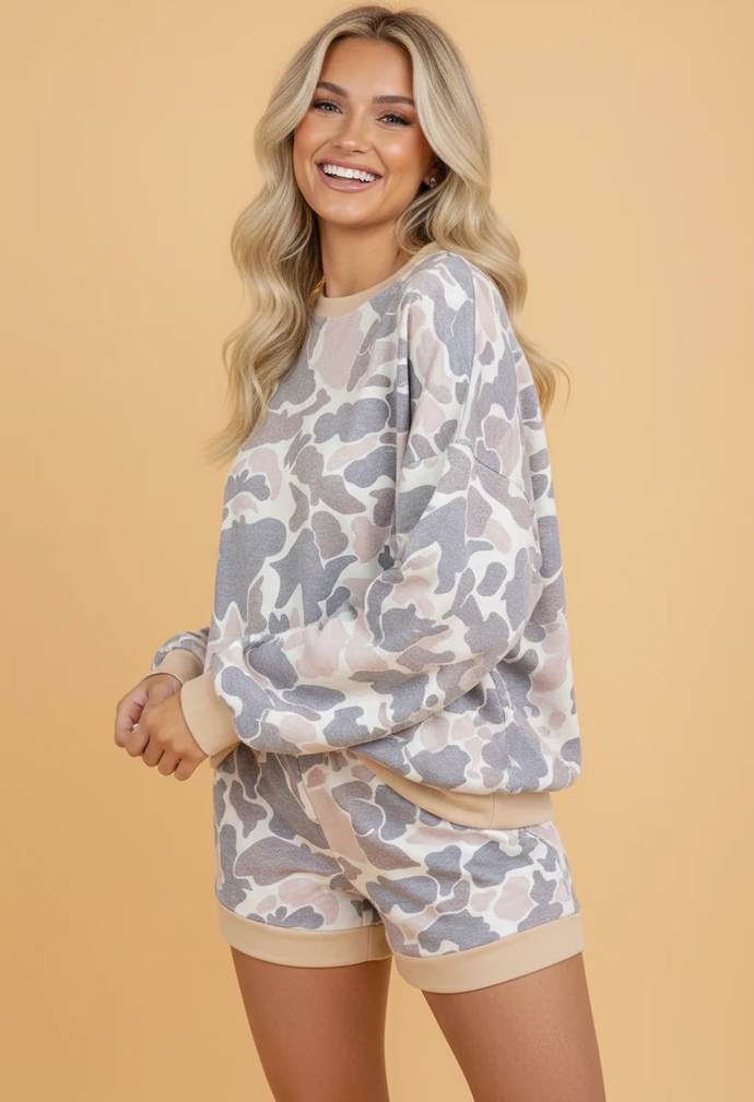 A smiling blonde woman models a matching lounge set featuring a long-sleeve sweatshirt and shorts in a gray and beige camouflage pattern with solid beige ribbed trim, set against a solid beige background.