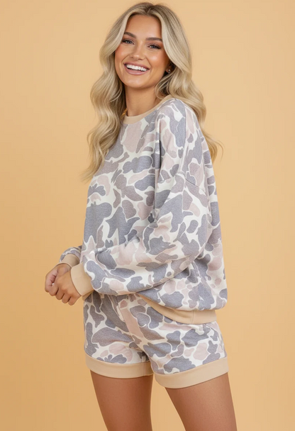 A smiling blonde woman models a matching lounge set featuring a long-sleeve sweatshirt and shorts in a gray and beige camouflage pattern with solid beige ribbed trim, set against a solid beige background.
