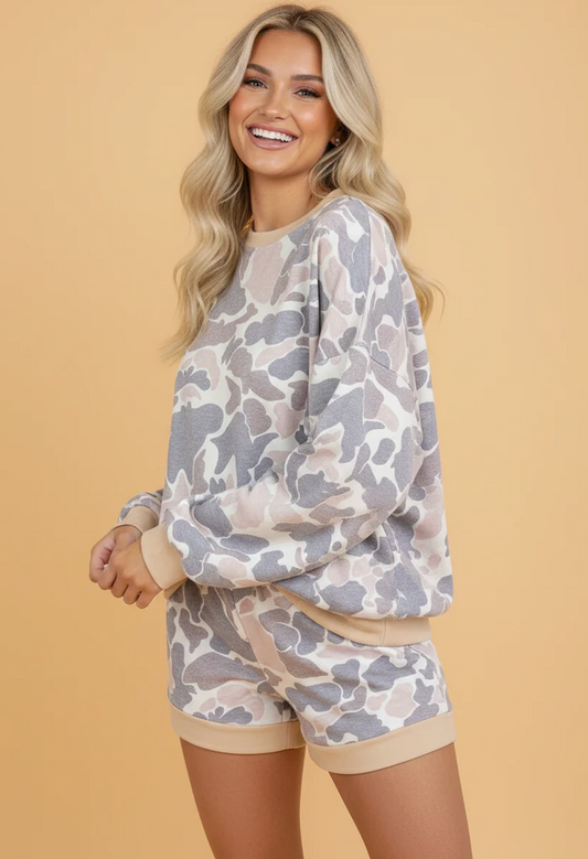 A smiling blonde woman models a matching lounge set featuring a long-sleeve sweatshirt and shorts in a gray and beige camouflage pattern with solid beige ribbed trim, set against a solid beige background.