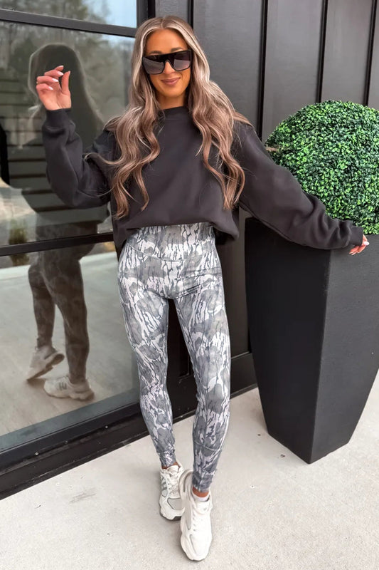 A woman wearing a dark charcoal gray sweatshirt and gray and white abstract patterned leggings, styled with white sneakers and black sunglasses, posing outdoors next to a black planter box.
