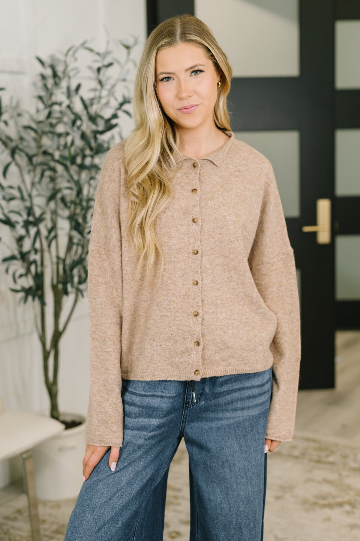 A woman modeling a beige button-up knit cardigan with a collar, paired with blue jeans, standing in a modern indoor setting with a potted plant and a black door in the background.