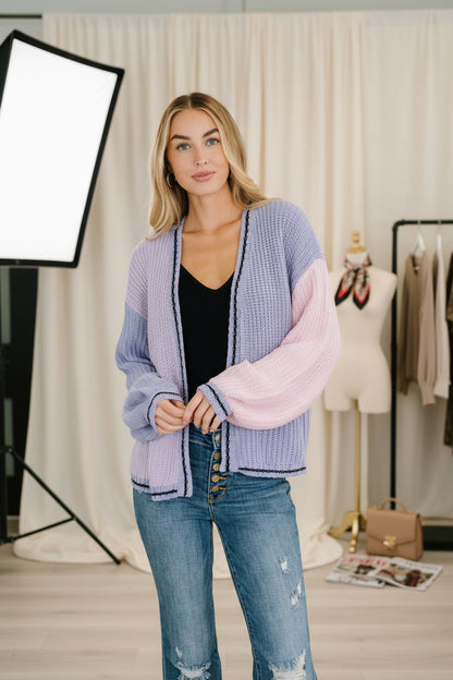 Woman wearing a colorful cardigan and jeans standing indoors with a plant in the background