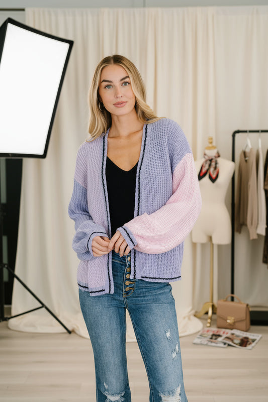 Woman wearing a colorful cardigan and jeans standing indoors with a plant in the background