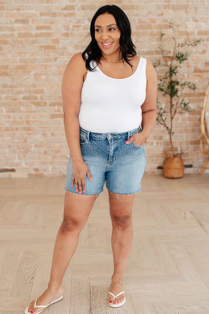Woman wearing a white tank top and denim shorts standing in front of a brick wall.