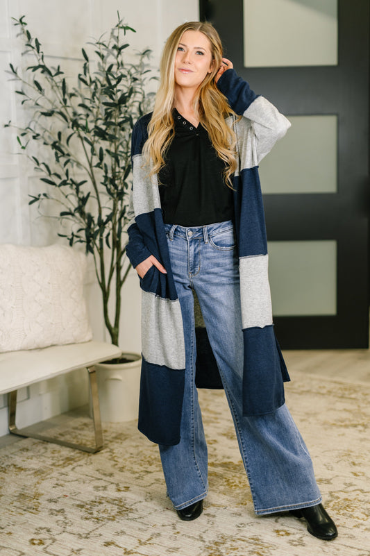 Woman wearing a long, patterned cardigan in a home setting with a plant and sofa.