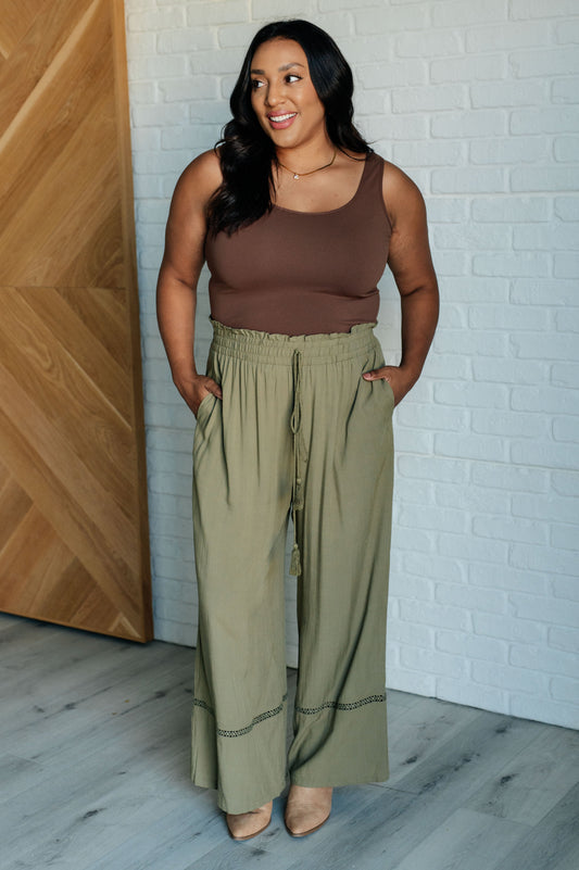 Woman wearing a brown tank top and green pants standing against a white brick wall.