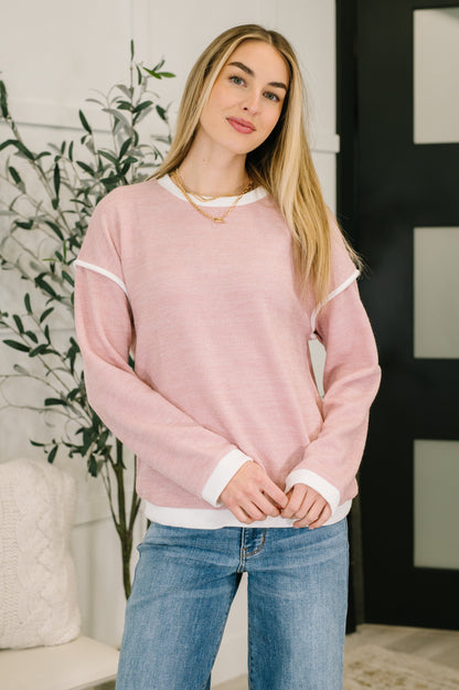 Woman wearing a pink sweater and blue jeans standing indoors with a plant in the background