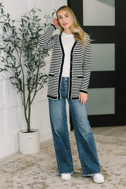 Woman wearing a black and white striped cardigan, white shirt, blue jeans, and white sneakers in an indoor setting.