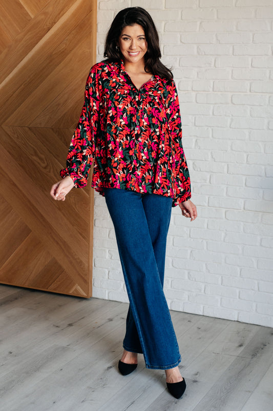 Woman wearing a colorful floral blouse and blue jeans standing against a white brick wall.