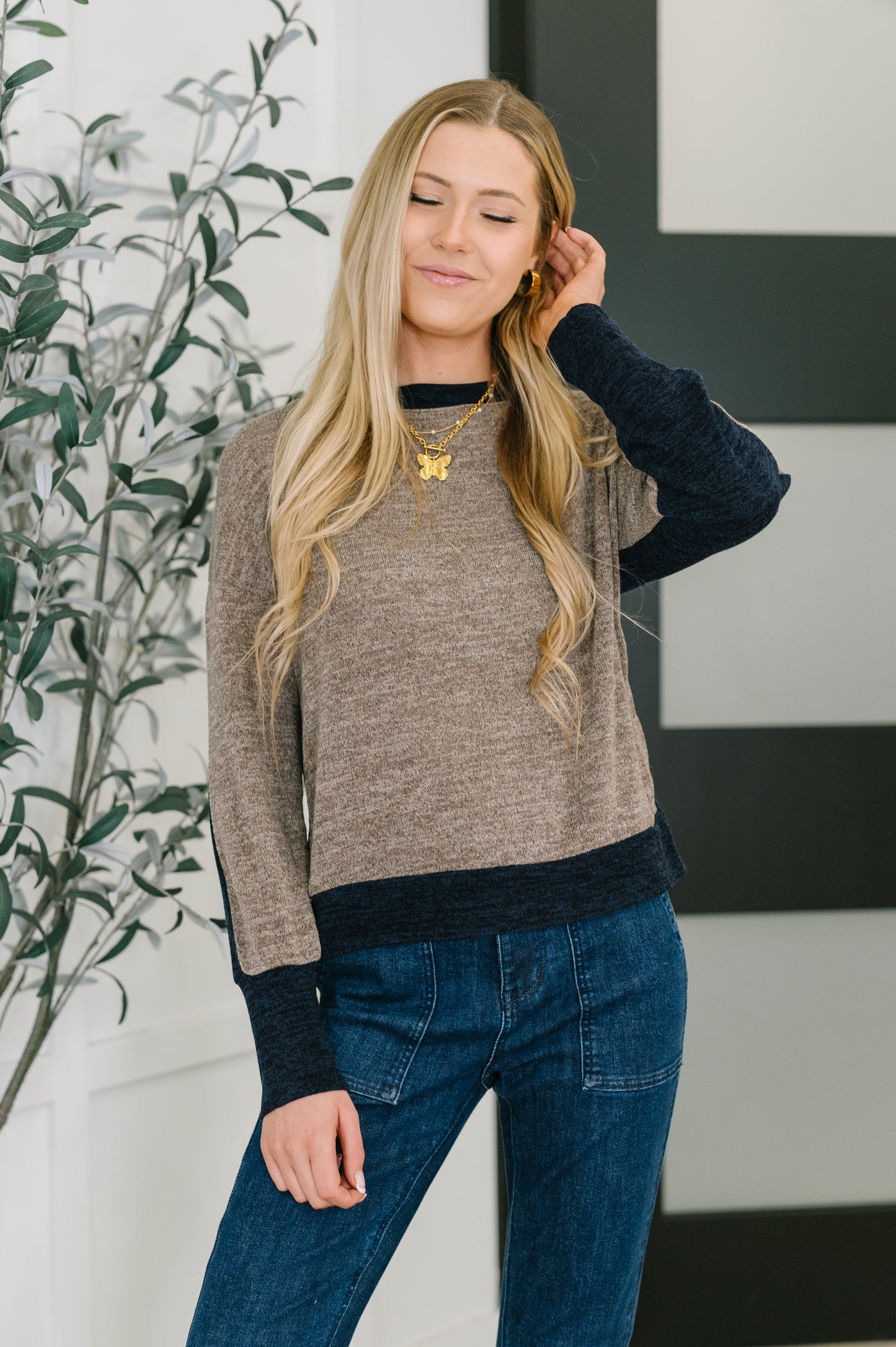 Woman wearing a brown sweater and blue jeans with a plant in the background