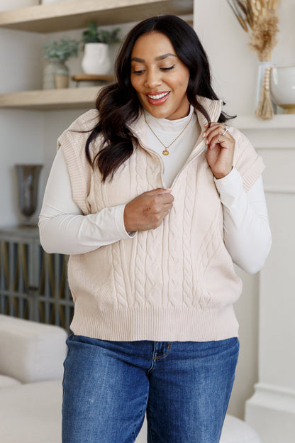 Woman wearing a beige cable knit vest over a white sweater and blue jeans in a cozy living room.