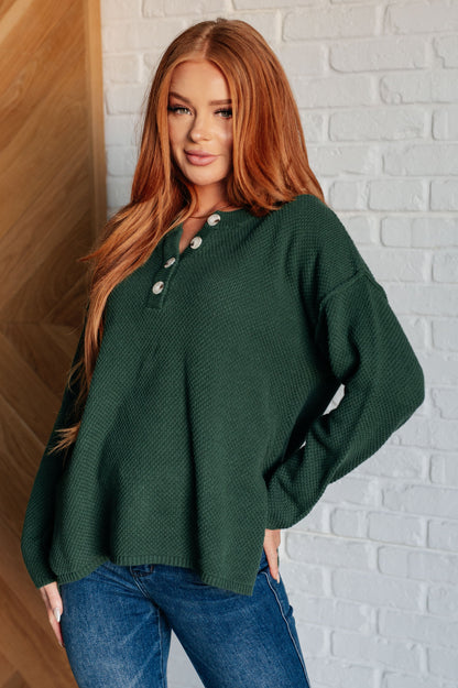 Woman wearing a green sweater with button details against a white brick wall.