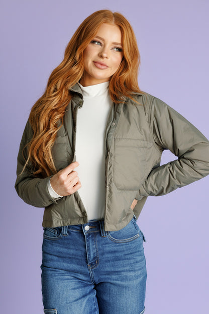 Woman wearing a green jacket over a white shirt and blue jeans against a white brick wall.