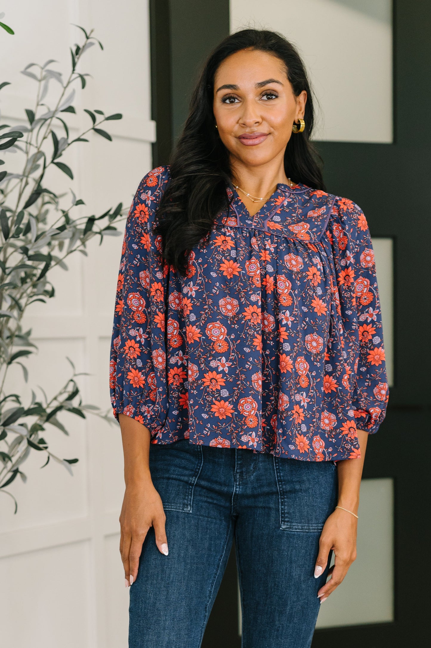 Woman wearing a floral blouse and jeans standing indoors with a plant in the background