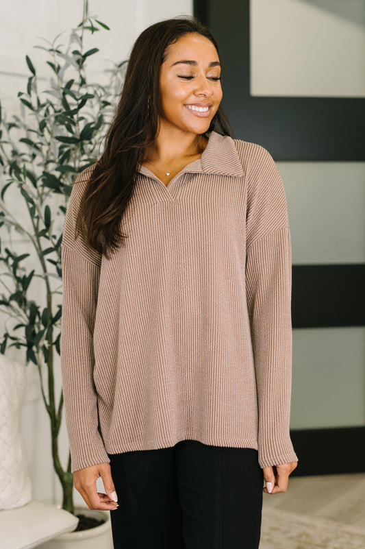 A woman wearing a ribbed knit long-sleeve polo-style sweater in a dusty rose color, paired with black pants, standing in a modern indoor setting with a plant in the background.