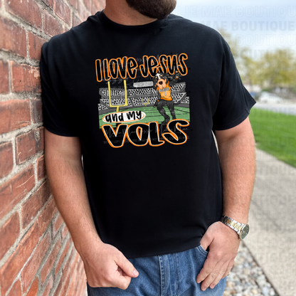 Man in black Gildan tee with “I Love Jesus and My Vols” design, leaning on brick wall, game-day ready.