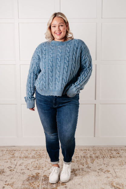 Woman wearing a blue cable knit sweater and jeans standing against a white paneled wall.