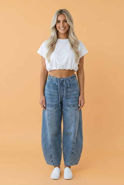 A woman modeling a white short-sleeve crop top and wide-leg blue denim jeans against a solid peach-colored studio background.
