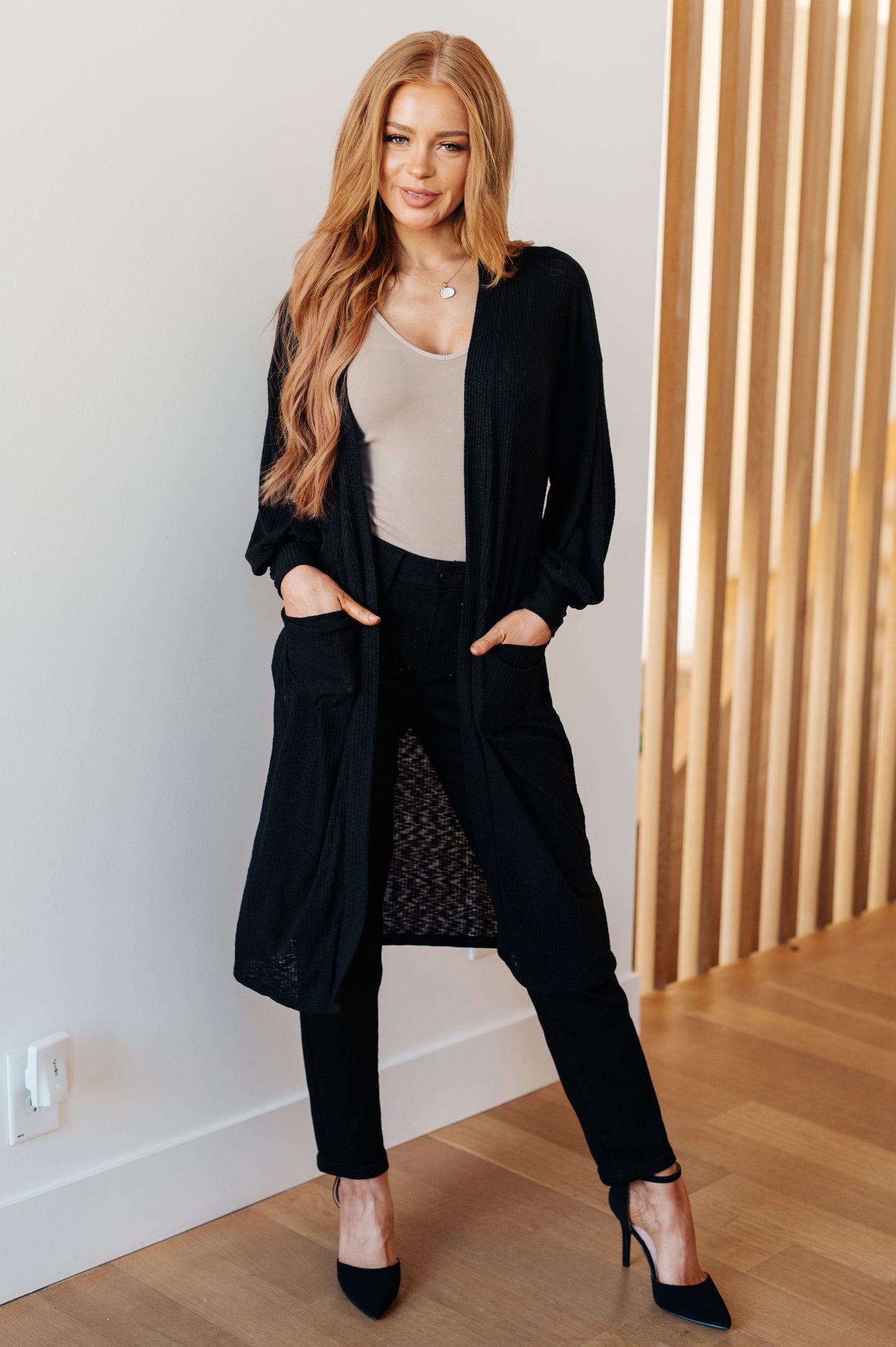 Woman wearing a black cardigan over a beige top and black pants in a minimal indoor setting.