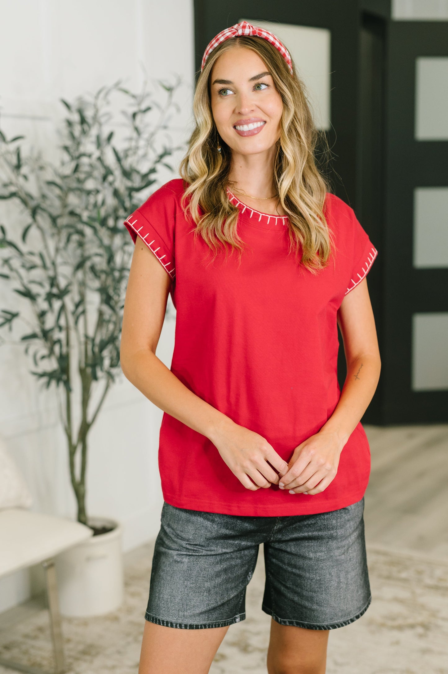 A woman wearing a bright red short-sleeve top with white contrast stitching along the neckline and sleeve hems, paired with gray denim shorts and a red-and-white patterned headband, standing in a modern indoor setting.