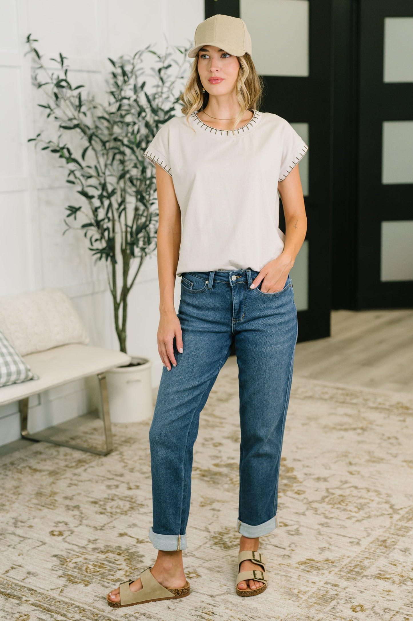 A woman stands indoors wearing a light beige short-sleeve top with decorative trim, blue cuffed jeans, a beige baseball cap, and tan sandals. The room has a light rug, a bench, and a potted plant.