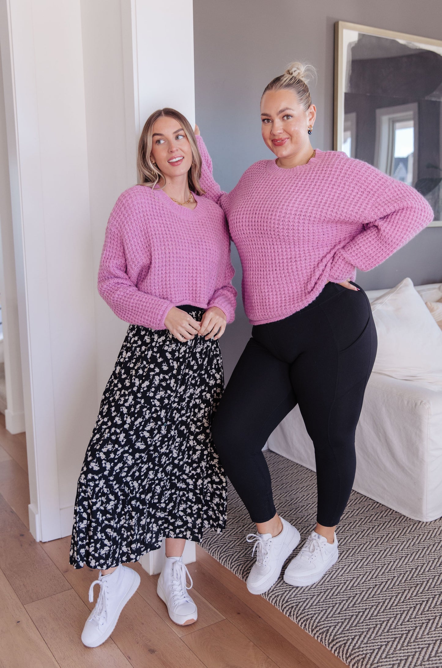 Two women wearing pink sweaters in a room with a mirror and bed in the background.