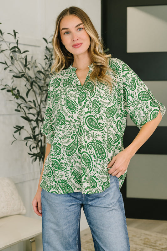 Woman wearing a green paisley blouse and blue jeans in a room with a plant and wall art.