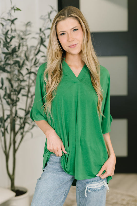 Woman wearing a green top and blue jeans standing indoors with a plant in the background