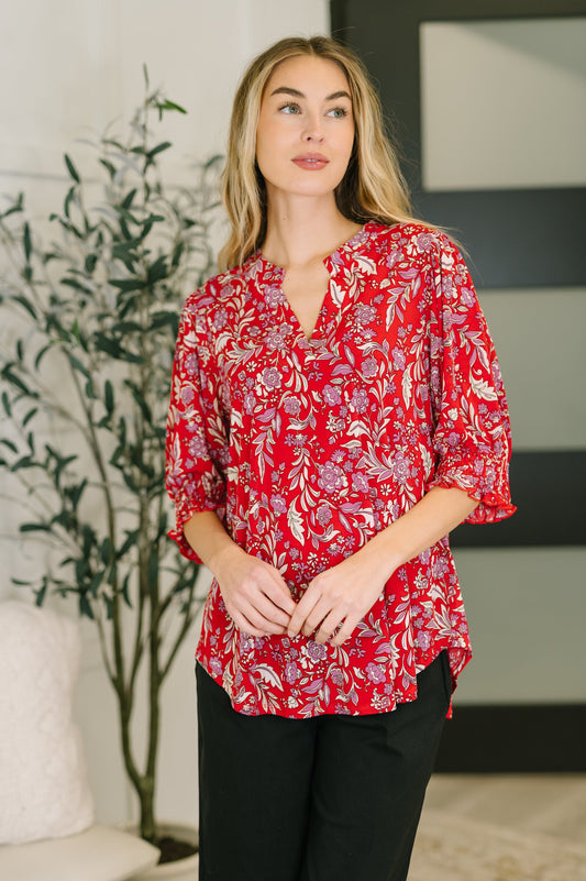 Woman wearing a red floral blouse standing in a room with a plant and framed picture.