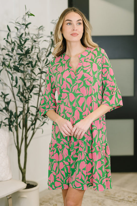 Woman wearing a pink and green patterned dress standing indoors with a plant in the background.