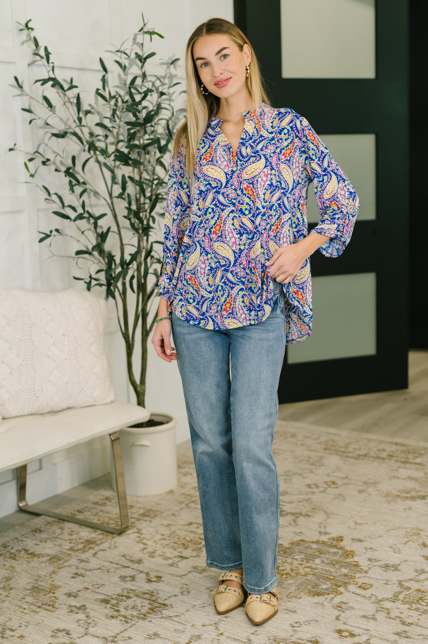 Woman wearing a colorful patterned blouse and blue jeans standing in a room with a plant and sofa.