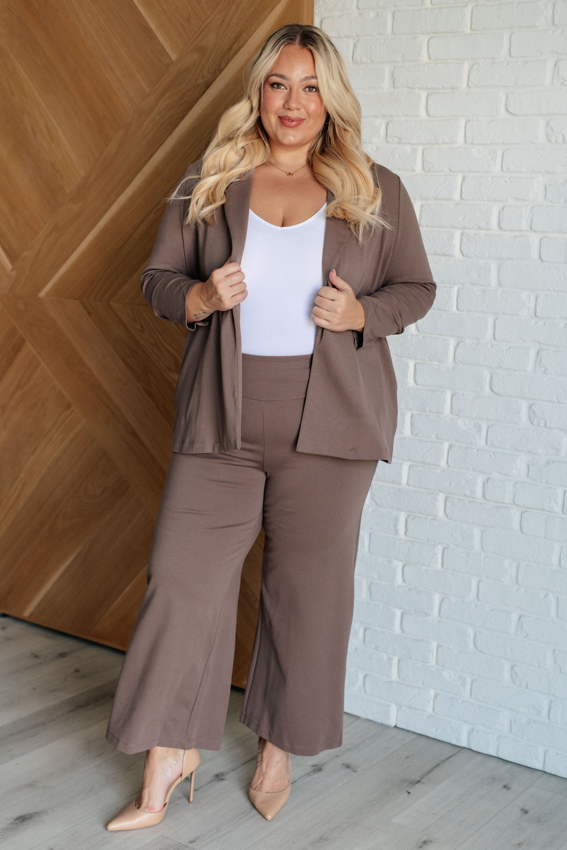 Woman wearing a brown suit standing against a white brick wall.
