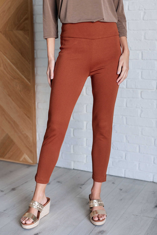 Person wearing rust-colored pants and a brown top against a white brick wall.