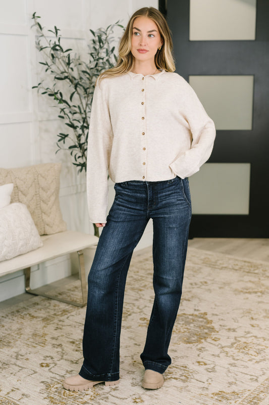 A woman modeling a cream-colored long-sleeve button-up cardigan and dark blue flare jeans in a modern, brightly lit living room setting.