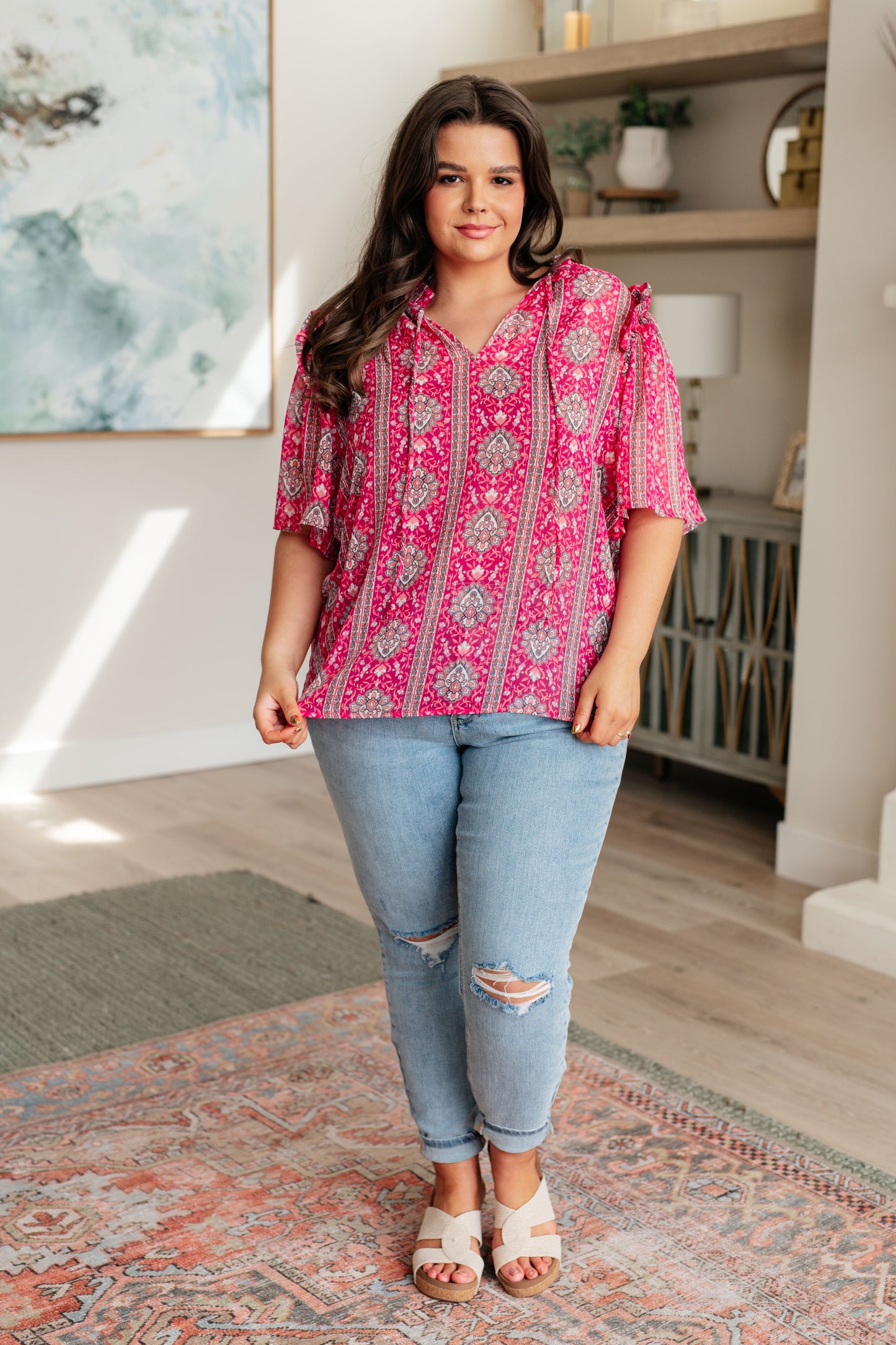 Woman wearing a pink patterned blouse and blue jeans standing in a room with a rug and shelves.