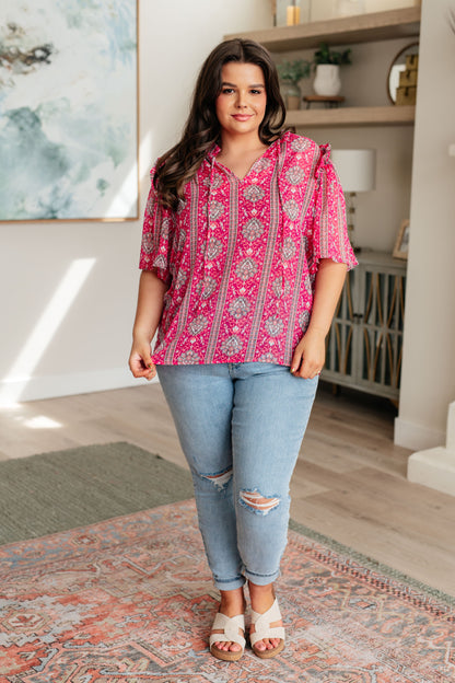 Woman wearing a pink patterned blouse and blue jeans standing in a room with a rug and shelves.