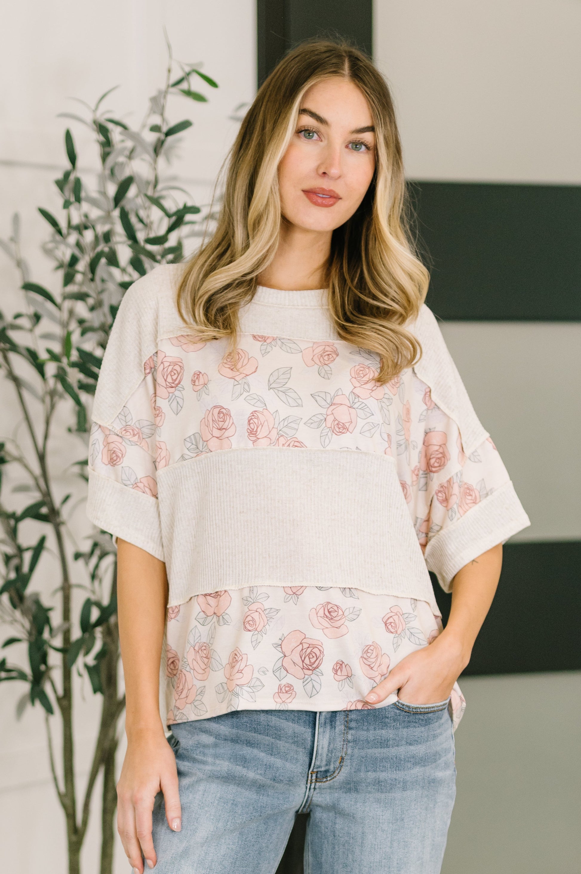 Woman wearing a floral blouse and jeans standing indoors with a plant in the background