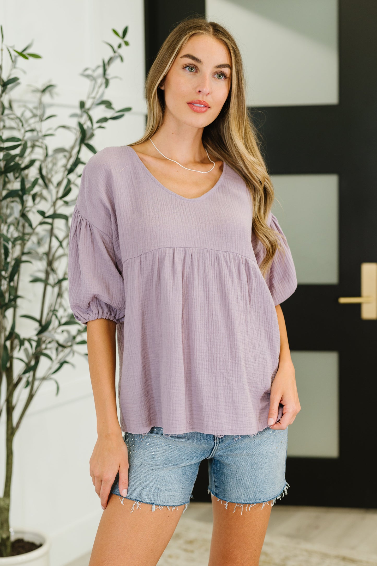 Woman wearing a light purple blouse and denim shorts standing indoors.
