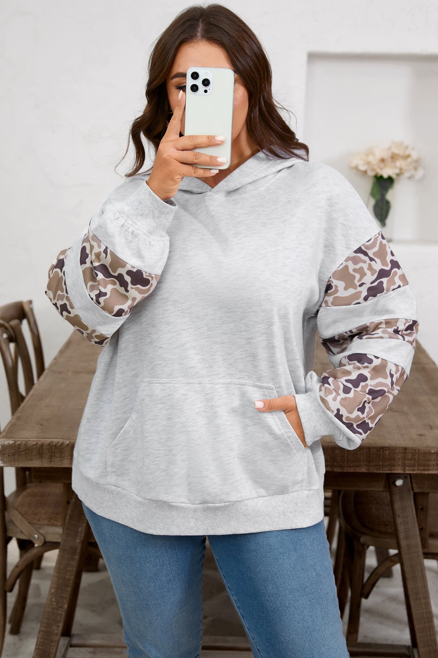Person wearing a gray hoodie with camouflage sleeves in a room with a wooden table and chairs.