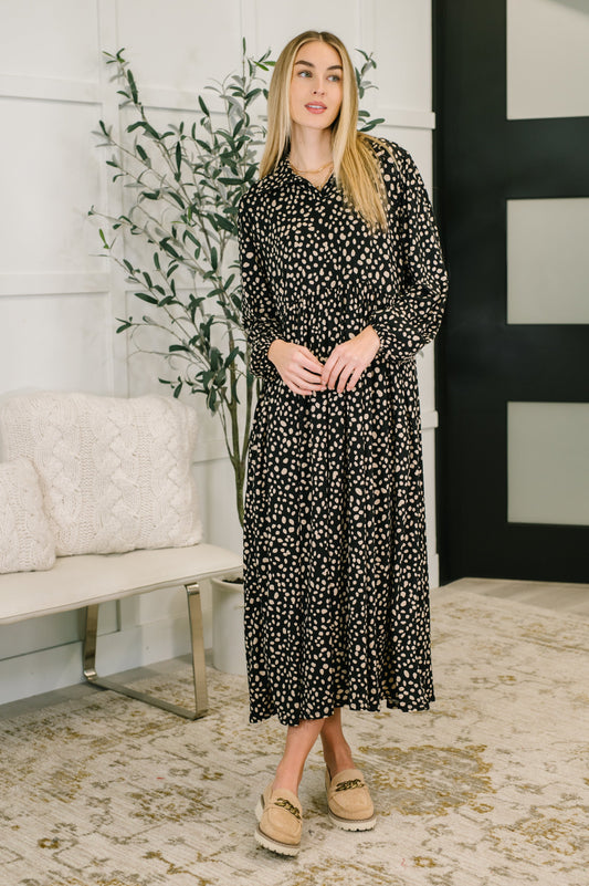 Woman wearing a black floral dress standing in a room with a white wall and decorative plant.