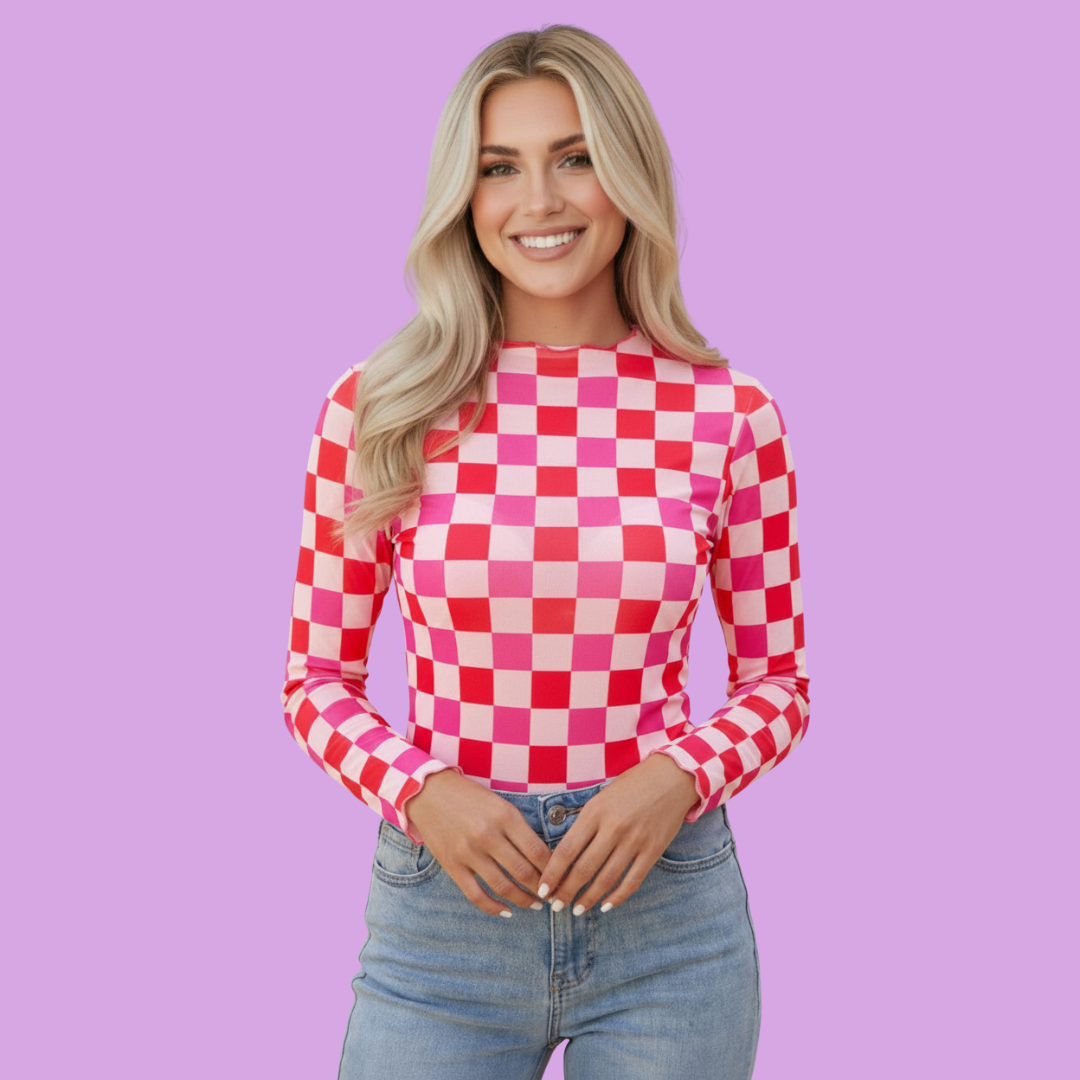 A smiling blonde woman wearing a pink and red checkerboard long-sleeve top with light blue jeans, standing against a solid lavender-purple background.