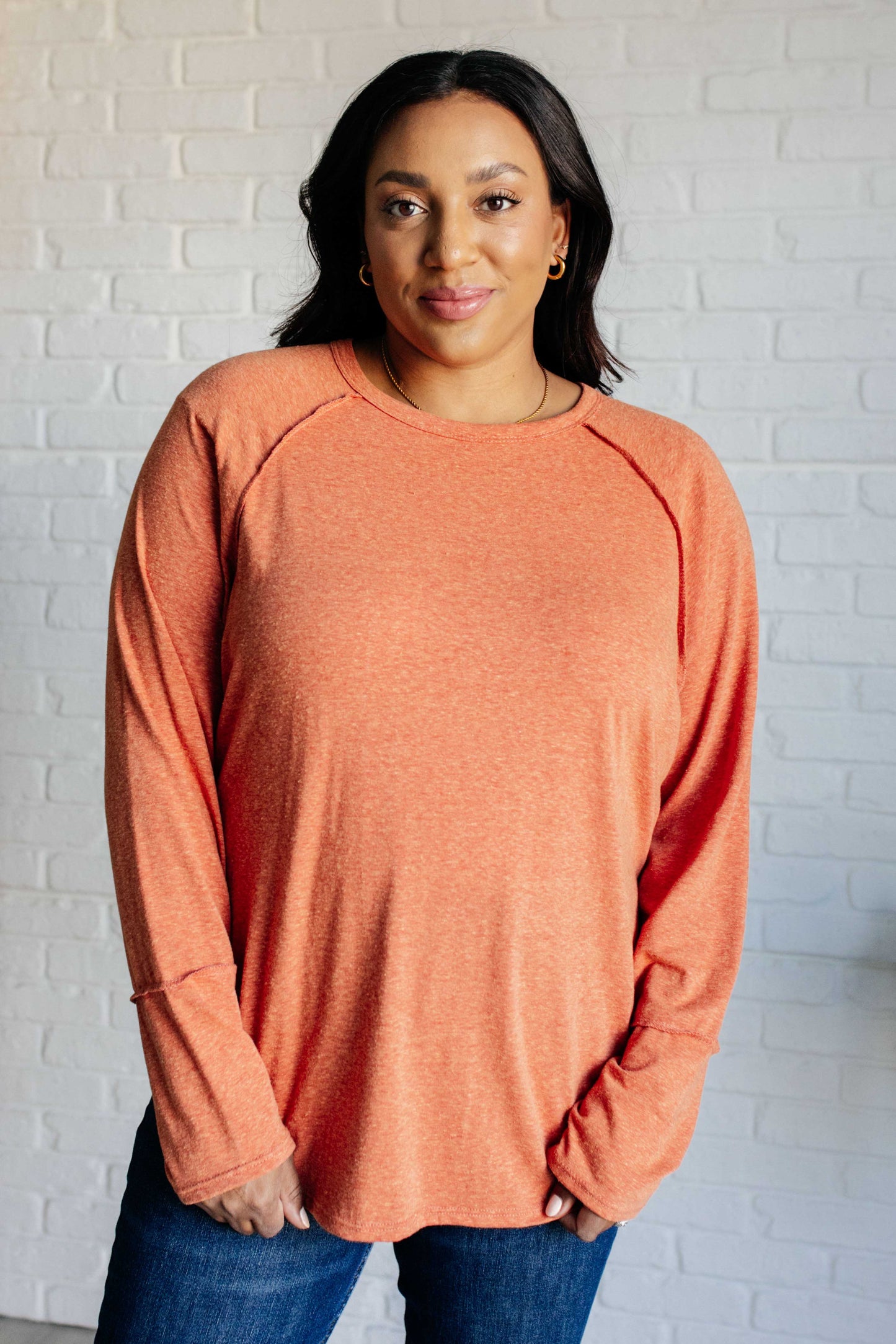 Woman wearing an orange long-sleeve shirt against a white brick wall.